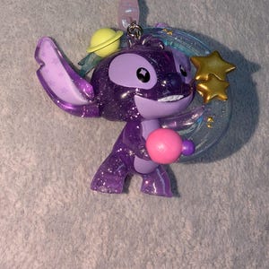 May include: A purple glittery Stitch keychain with a planet, stars, and a pink ball. The keychain has a silver chain and a light purple bead. The Stitch figure is holding a pink ball and has a yellow planet on its head.