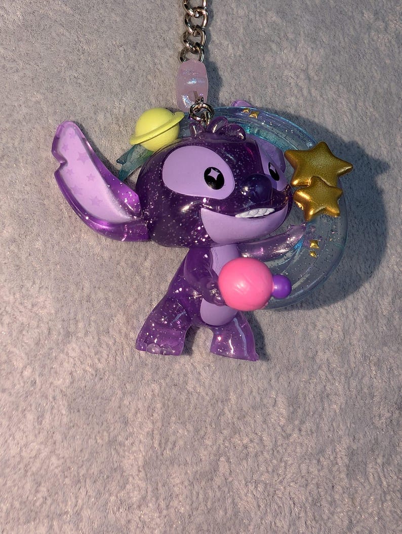 May include: A purple glittery Stitch keychain with a planet, stars, and a pink ball. The keychain has a silver chain and a light purple bead. The Stitch figure is holding a pink ball and has a yellow planet on its head.