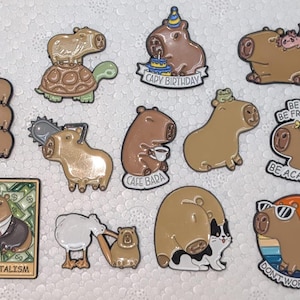 Puede incluir: Una colección de pines de esmalte de carpincho en varios diseños. Algunos pines presentan carpinchos con accesorios como una motosierra, gafas de sol o un gorro de cumpleaños. Otros pines tienen texto que dice "CAPY BIRTHDAY", "CAFE BARA", "CAPY-ITALISM" y "DON'T WORRY BE CAPY".