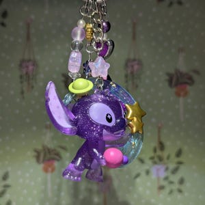 May include: A purple glittery Stitch keychain with a pink ball, a green planet, and a gold star. The keychain has a silver ring and is adorned with purple, pink, and gold beads. The background is a light green wallpaper with hanging plant designs.