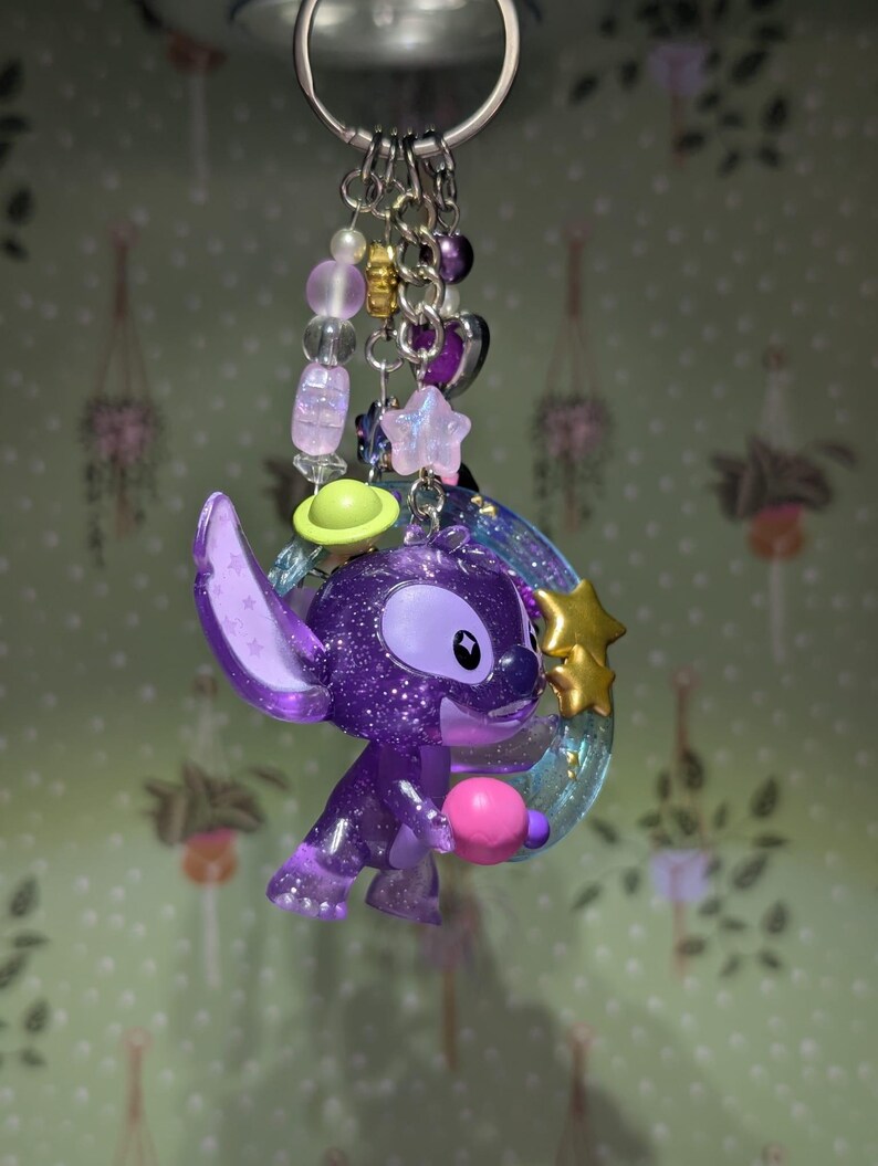 May include: A purple glittery Stitch keychain with a pink ball, a green planet, and a gold star. The keychain has a silver ring and is adorned with purple, pink, and gold beads. The background is a light green wallpaper with hanging plant designs.