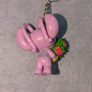 May include: Pink keychain featuring a cartoon character holding a bouquet of flowers. The character has a rounded body, long arms, and a small head. The flowers are red and green. The keychain has a silver ring.