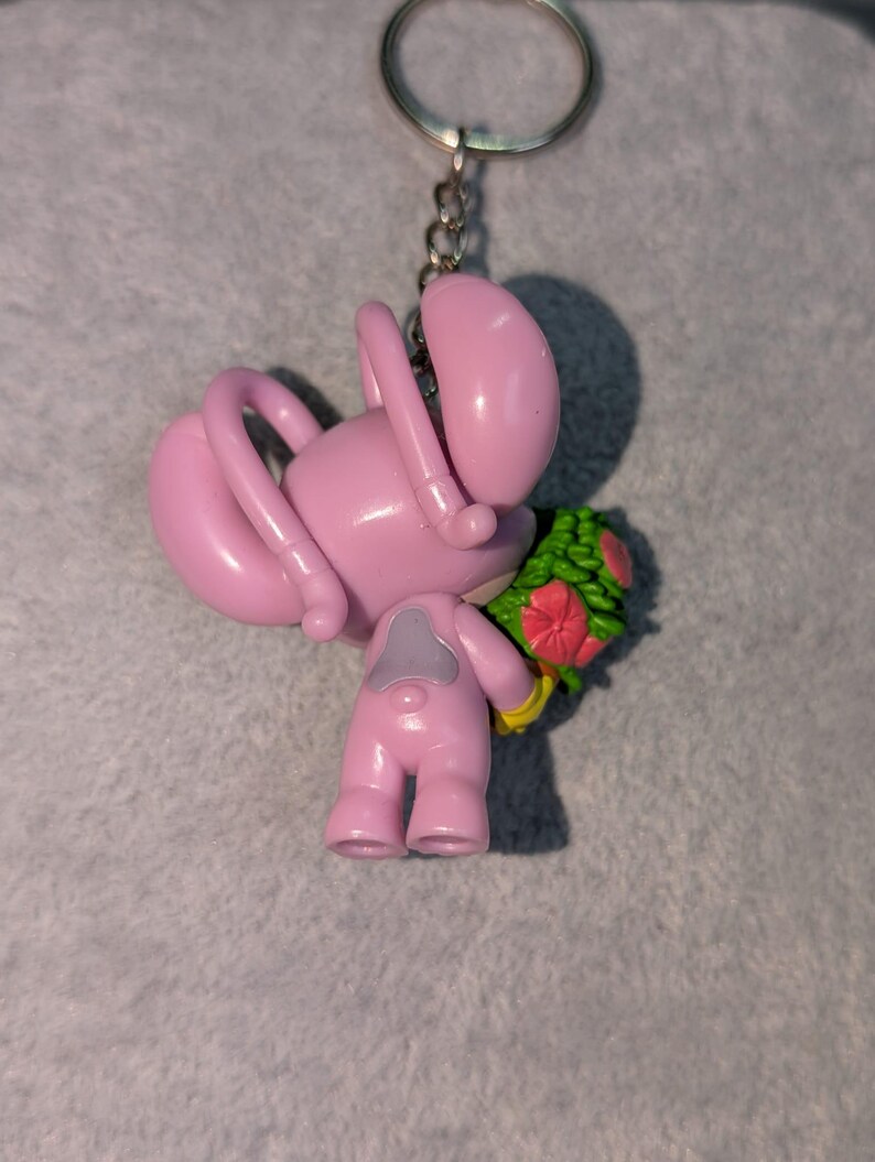 May include: Pink keychain featuring a cartoon character holding a bouquet of flowers. The character has a rounded body, long arms, and a small head. The flowers are red and green. The keychain has a silver ring.