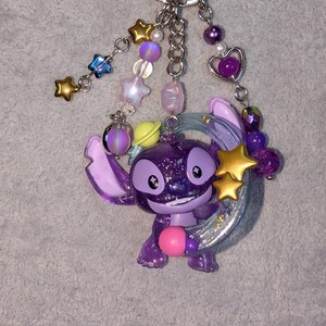 May include: A purple glittery Stitch keychain with a pink ball, gold stars, and a silver crescent moon. The keychain has a silver ring and is adorned with various beads in shades of purple, pink, and blue.