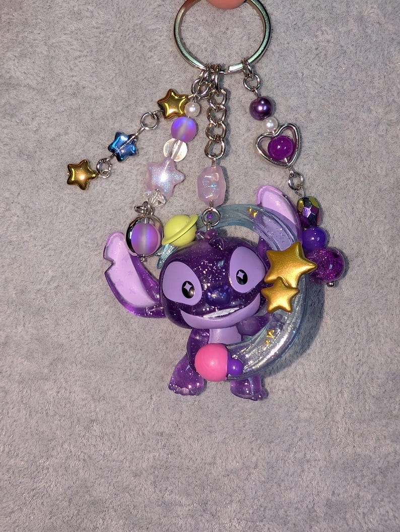 May include: A purple glittery Stitch keychain with a pink ball, gold stars, and a silver crescent moon. The keychain has a silver ring and is adorned with various beads in shades of purple, pink, and blue.