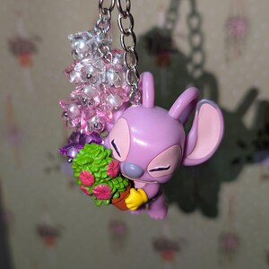 May include: A lavender-colored Angel keychain with a silver chain and a cluster of pink and clear beads. Angel is holding a small potted plant with green leaves and pink flowers. The keychain is attached to a silver keyring.