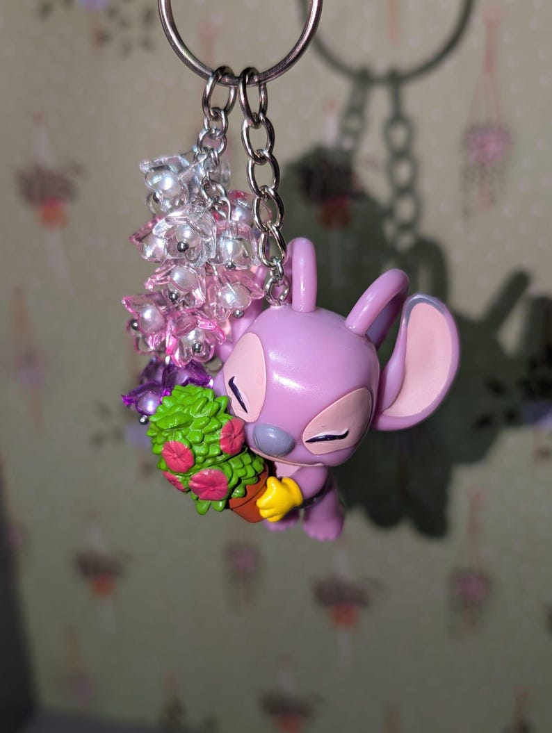 May include: A lavender-colored Angel keychain with a silver chain and a cluster of pink and clear beads. Angel is holding a small potted plant with green leaves and pink flowers. The keychain is attached to a silver keyring.
