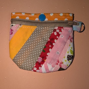 May include: A small, quilted coin purse with a patchwork design. The purse features a variety of fabrics in orange, yellow, pink, red, and gray, with polka dots and floral patterns. It has a zipper closure, a blue button, and a small metal ring.