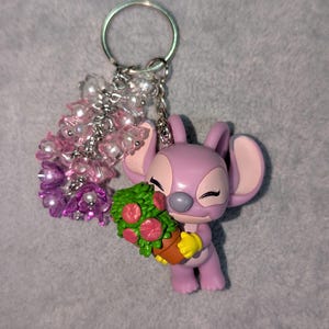 May include: A lavender Angel keychain holding a bouquet of flowers. The keychain features a silver ring and a cluster of pink and white beads. The flowers are red and green, and the figure is wearing yellow gloves.