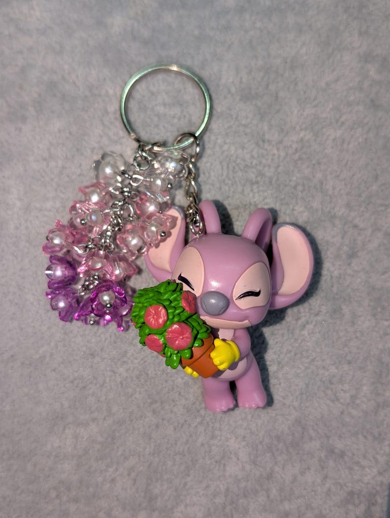 May include: A lavender Angel keychain holding a bouquet of flowers. The keychain features a silver ring and a cluster of pink and white beads. The flowers are red and green, and the figure is wearing yellow gloves.