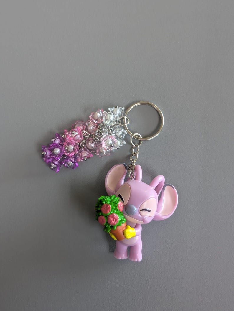 May include: A keychain featuring a purple character holding a green and pink floral arrangement. The keychain also includes a beaded floral charm in shades of pink, purple, and silver. The background is a solid gray.