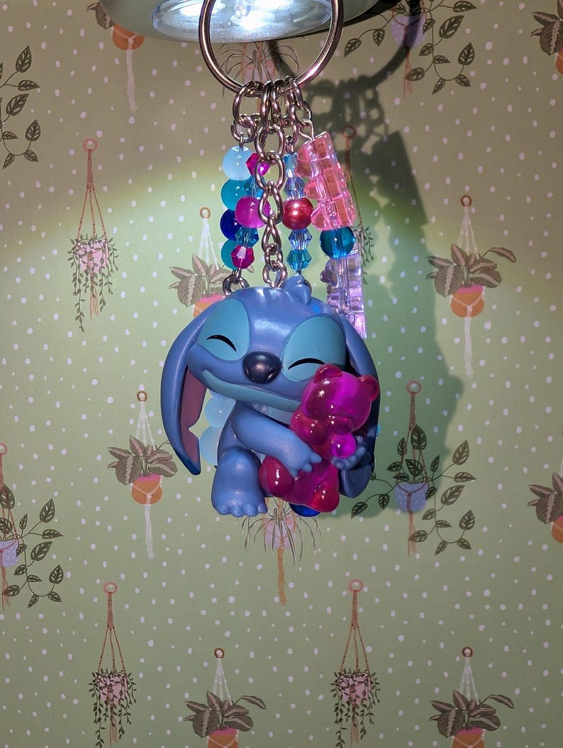 May include: A blue Stitch keychain holding a pink teddy bear. The keychain has a silver ring and is adorned with blue, pink, and clear beads. The background is a green wallpaper with hanging plant illustrations.