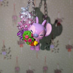 May include: A keychain featuring a lavender-colored cartoon character holding a bouquet of flowers in a small pot. The keychain includes a silver chain and a cluster of pink and clear beads. The background is a light green wallpaper with a floral pattern.