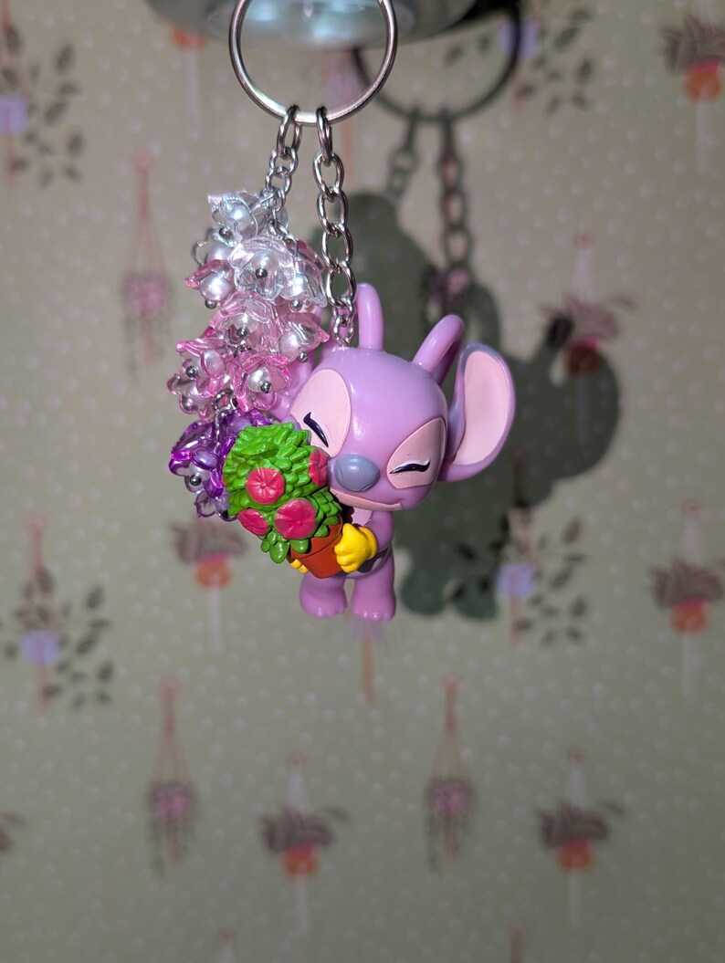 May include: A keychain featuring a lavender-colored cartoon character holding a bouquet of flowers in a small pot. The keychain includes a silver chain and a cluster of pink and clear beads. The background is a light green wallpaper with a floral pattern.