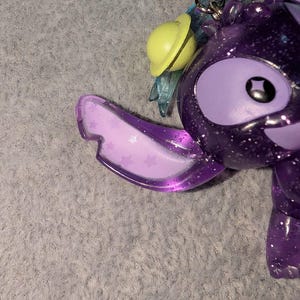 May include: A purple glittery character keychain with large eyes and a smiling expression. The keychain has a yellow planet-shaped charm and translucent blue wings. The character's ear is a translucent purple with star-shaped designs.