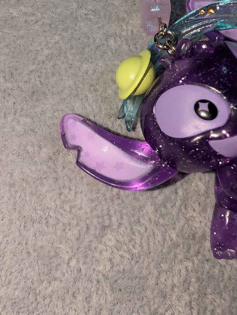 May include: A purple glittery character keychain with large eyes and a smiling expression. The keychain has a yellow planet-shaped charm and translucent blue wings. The character's ear is a translucent purple with star-shaped designs.