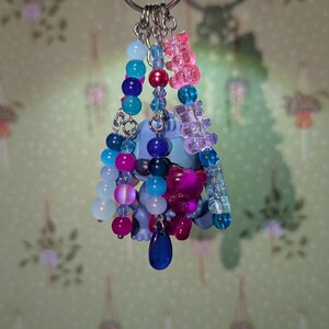 May include: A handmade keychain featuring a blue cartoon character and various colorful beads. The keychain includes pink, blue, and clear beads, along with a dark blue teardrop charm. The beads are strung on silver-colored chains.