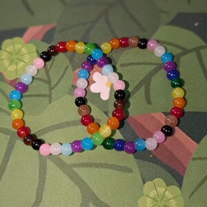 Könnte beinhalten: Zwei Armbänder mit Perlen in Regenbogenfarben. Die Perlen sind kreisförmig angeordnet, mit Farben wie Rot, Orange, Gelb, Grün, Blau und Lila. Die Armbänder liegen auf einem grünen Hintergrund.