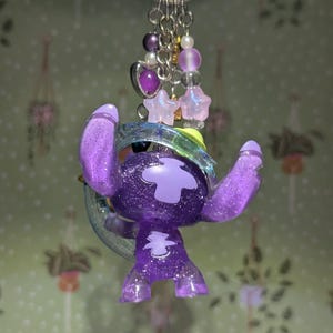 May include: A purple glittery Stitch keychain with a silver keyring. The Stitch figure has a large head, big ears, and a cross-shaped design on its chest. The keychain also features a cluster of purple beads, stars, and a heart charm.