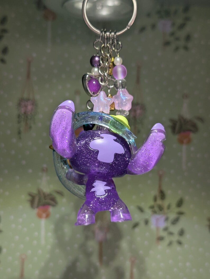 May include: A purple glittery Stitch keychain with a silver keyring. The Stitch figure has a large head, big ears, and a cross-shaped design on its chest. The keychain also features a cluster of purple beads, stars, and a heart charm.
