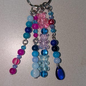 May include: A silver keychain with multiple beaded charms in shades of blue, pink, and purple. The charms include round beads, faceted crystals, and gummy bear shapes. A dark blue teardrop-shaped charm hangs at the end of one strand.