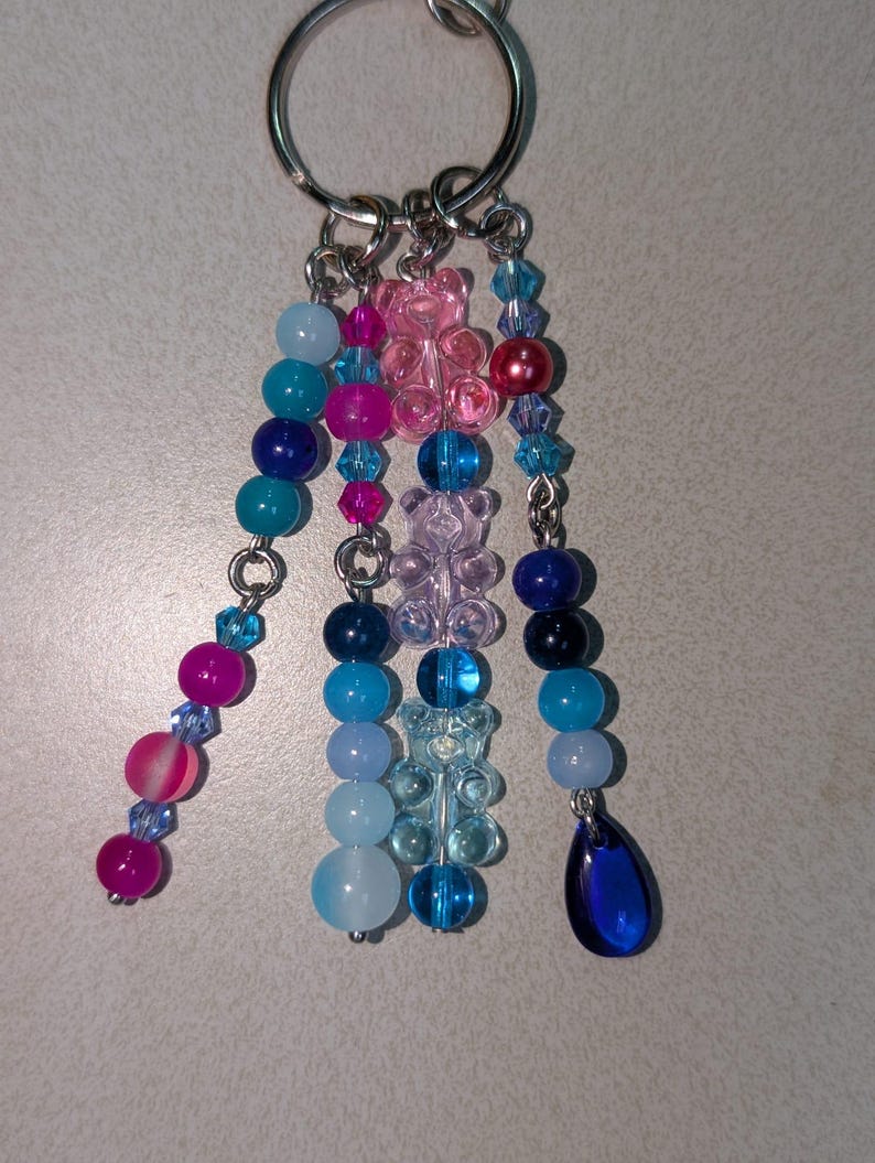 May include: A silver keychain with multiple beaded charms in shades of blue, pink, and purple. The charms include round beads, faceted crystals, and gummy bear shapes. A dark blue teardrop-shaped charm hangs at the end of one strand.