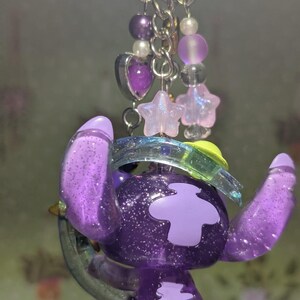 May include: A purple glittery keychain featuring a cartoon character with large ears and a light purple patch. The keychain includes a silver ring, purple beads, and star-shaped charms. The character is holding a clear, green-tinted object.
