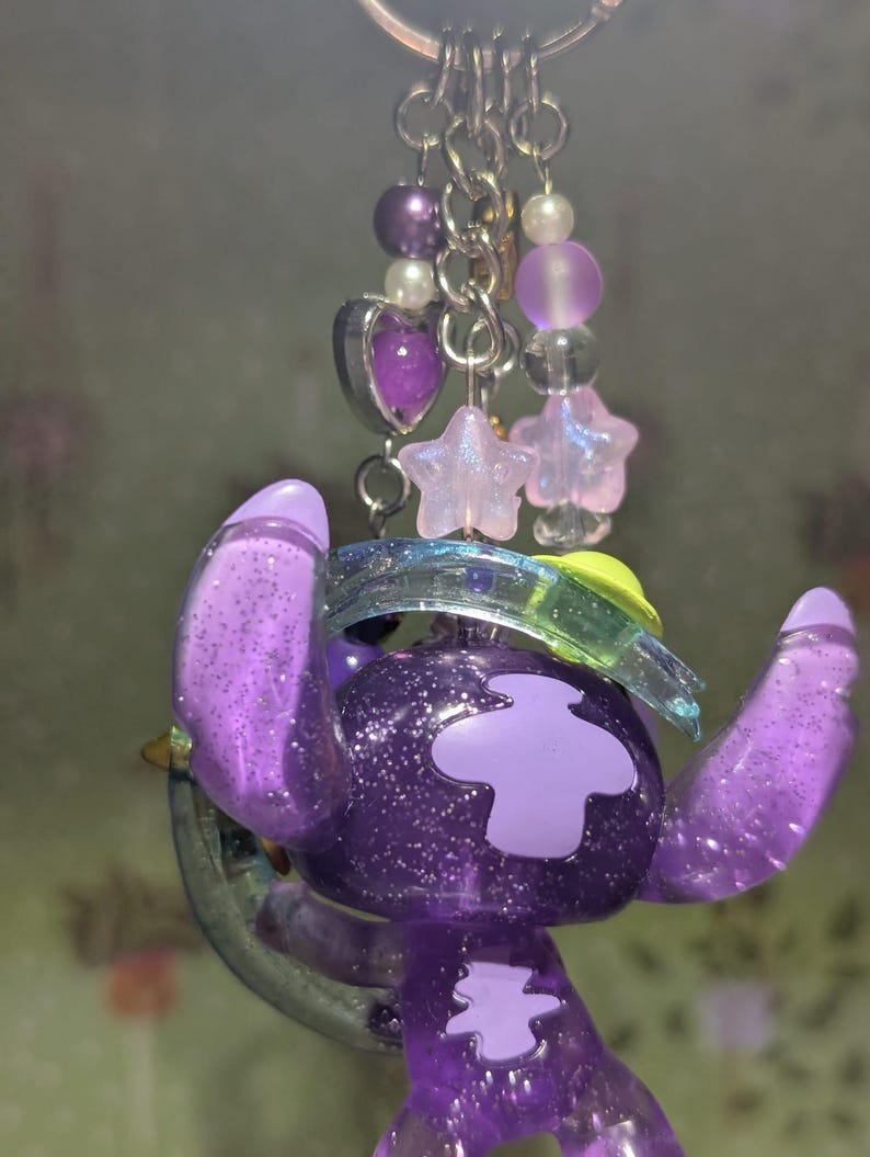 May include: A purple glittery keychain featuring a cartoon character with large ears and a light purple patch. The keychain includes a silver ring, purple beads, and star-shaped charms. The character is holding a clear, green-tinted object.