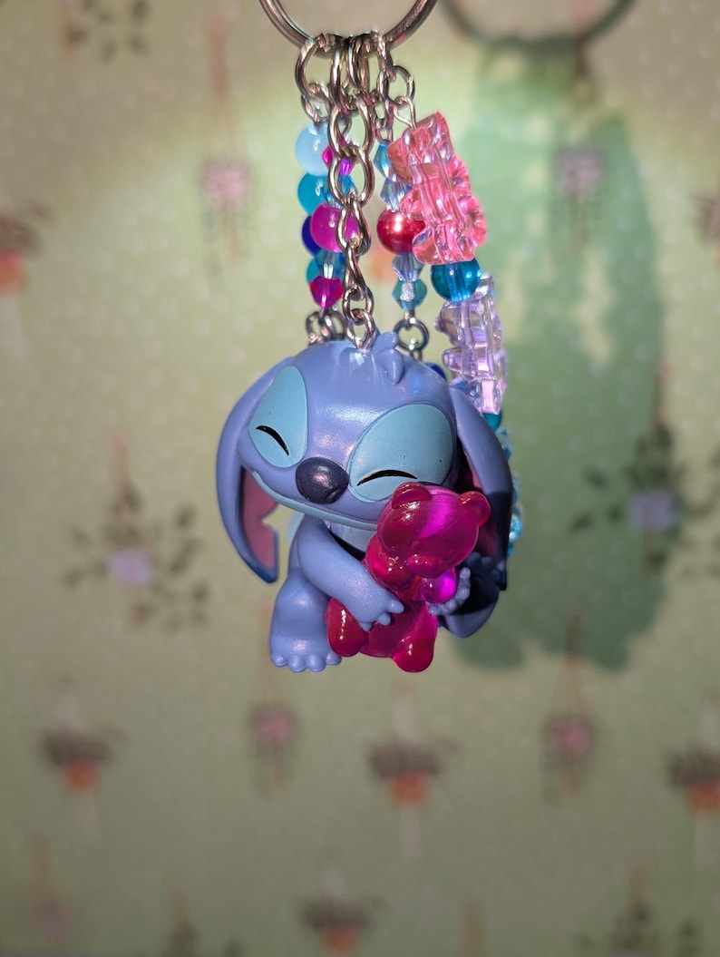 May include: A keychain featuring a blue Stitch figurine holding a pink teddy bear. The keychain has a silver ring and is adorned with colorful beads in shades of pink, blue, and purple. A charming accessory.