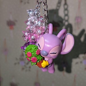 May include: A lavender-colored keychain featuring a cartoon character holding a small potted plant with pink flowers. The keychain includes a silver chain and a cluster of clear and pink beads. The character has large ears and yellow gloves.