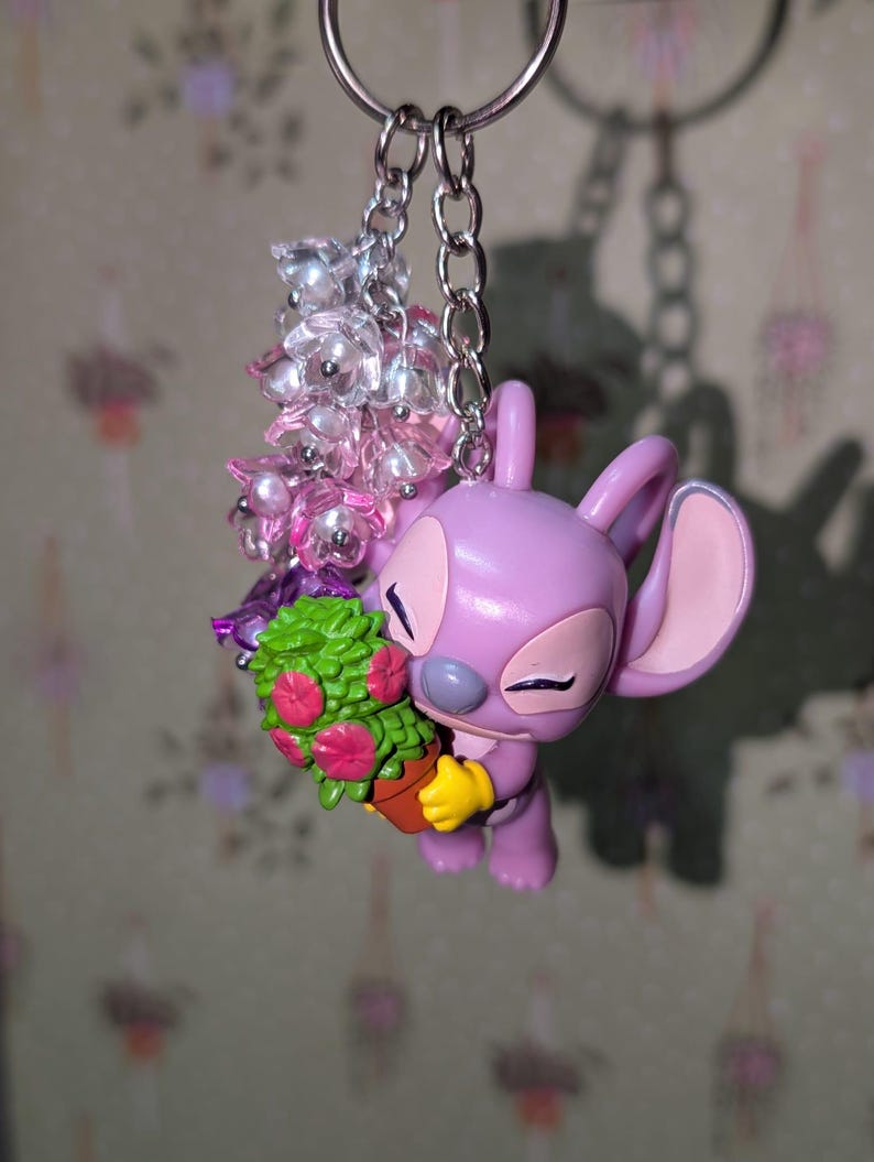 May include: A lavender-colored keychain featuring a cartoon character holding a small potted plant with pink flowers. The keychain includes a silver chain and a cluster of clear and pink beads. The character has large ears and yellow gloves.