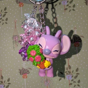 May include: A lavender-colored Angel keychain holding a potted plant with pink flowers and green leaves. The keychain also features a cluster of clear and pink beads. The keychain is attached to a silver metal ring.