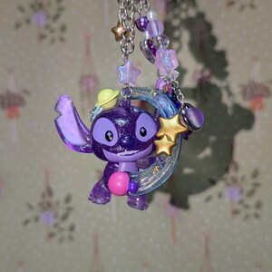 May include: A purple Stitch keychain with a glittery finish, holding a pink heart. The keychain features a silver chain with star and heart charms, and purple and white beads. A yellow hat is on Stitch's head.