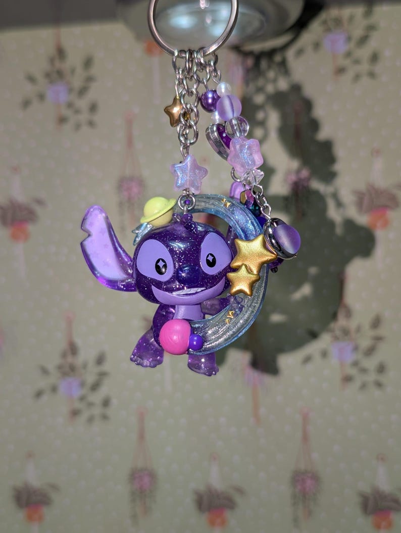May include: A purple Stitch keychain with a glittery finish, holding a pink heart. The keychain features a silver chain with star and heart charms, and purple and white beads. A yellow hat is on Stitch's head.