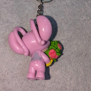 May include: Pink keychain featuring a cartoon figure holding a bouquet of flowers. The figure has a large head, curved arms, and small legs. The bouquet has green leaves and pink and red flowers. The keychain has a silver ring.