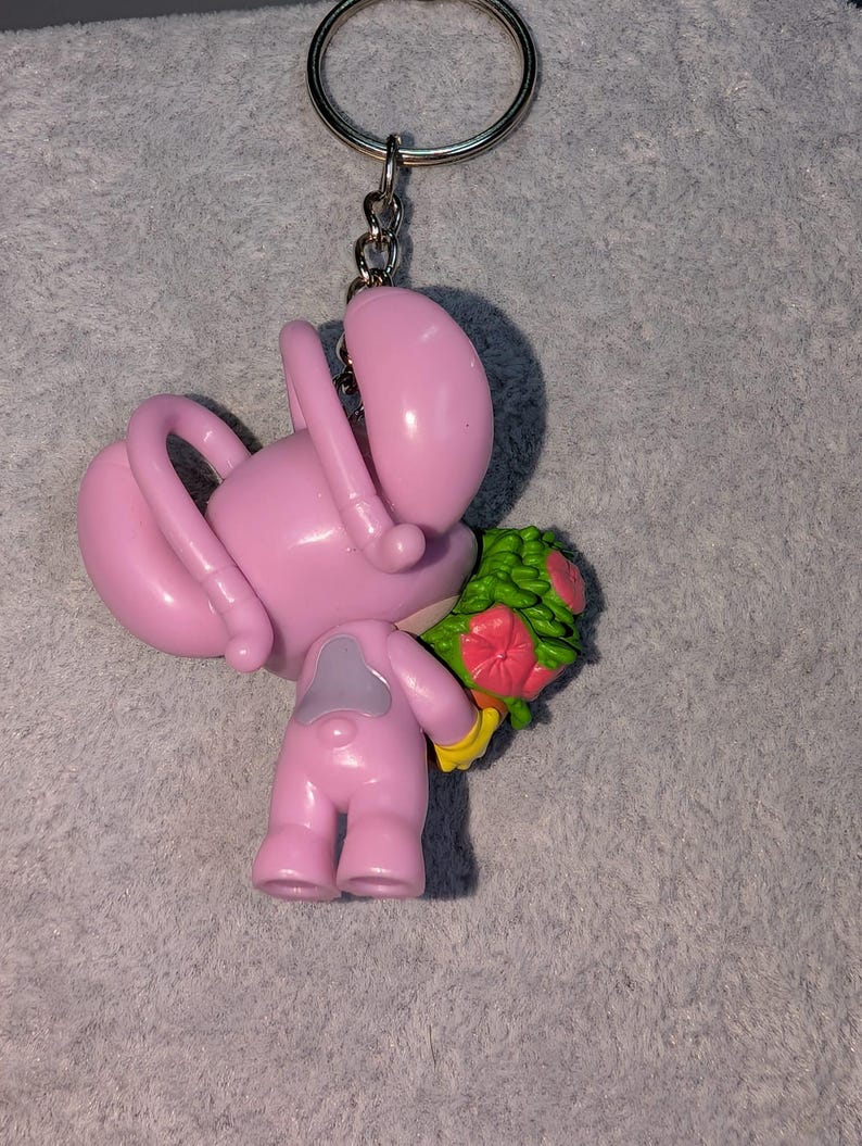 May include: Pink keychain featuring a cartoon figure holding a bouquet of flowers. The figure has a large head, curved arms, and small legs. The bouquet has green leaves and pink and red flowers. The keychain has a silver ring.