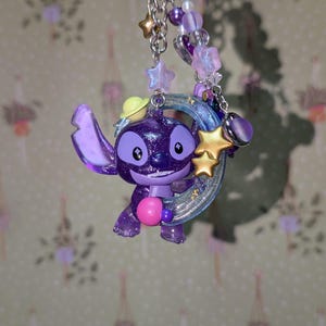 May include: A purple glittery Stitch keychain with a yellow hat, holding a pink ball. The keychain features a silver chain, star and bead accents, and a crescent moon. The charm is approximately 2 inches tall.