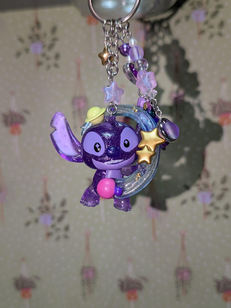 May include: A purple glittery Stitch keychain with a yellow hat, holding a pink ball. The keychain features a silver chain, star and bead accents, and a crescent moon. The charm is approximately 2 inches tall.