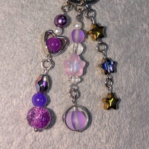 May include: A silver keychain with multiple charms. Charms include purple and iridescent beads, a heart-shaped charm, star-shaped charms in gold and blue, and a round charm with a purple orb inside. The keychain is on a gray surface.
