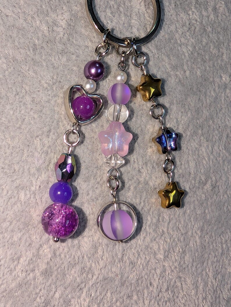 May include: A silver keychain with multiple charms. Charms include purple and iridescent beads, a heart-shaped charm, star-shaped charms in gold and blue, and a round charm with a purple orb inside. The keychain is on a gray surface.