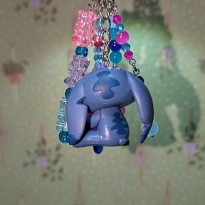 May include: A blue Stitch keychain with a silver keyring. The keychain features a Stitch figurine and colorful beads in pink, blue, and purple. The beads are strung together with silver chains.