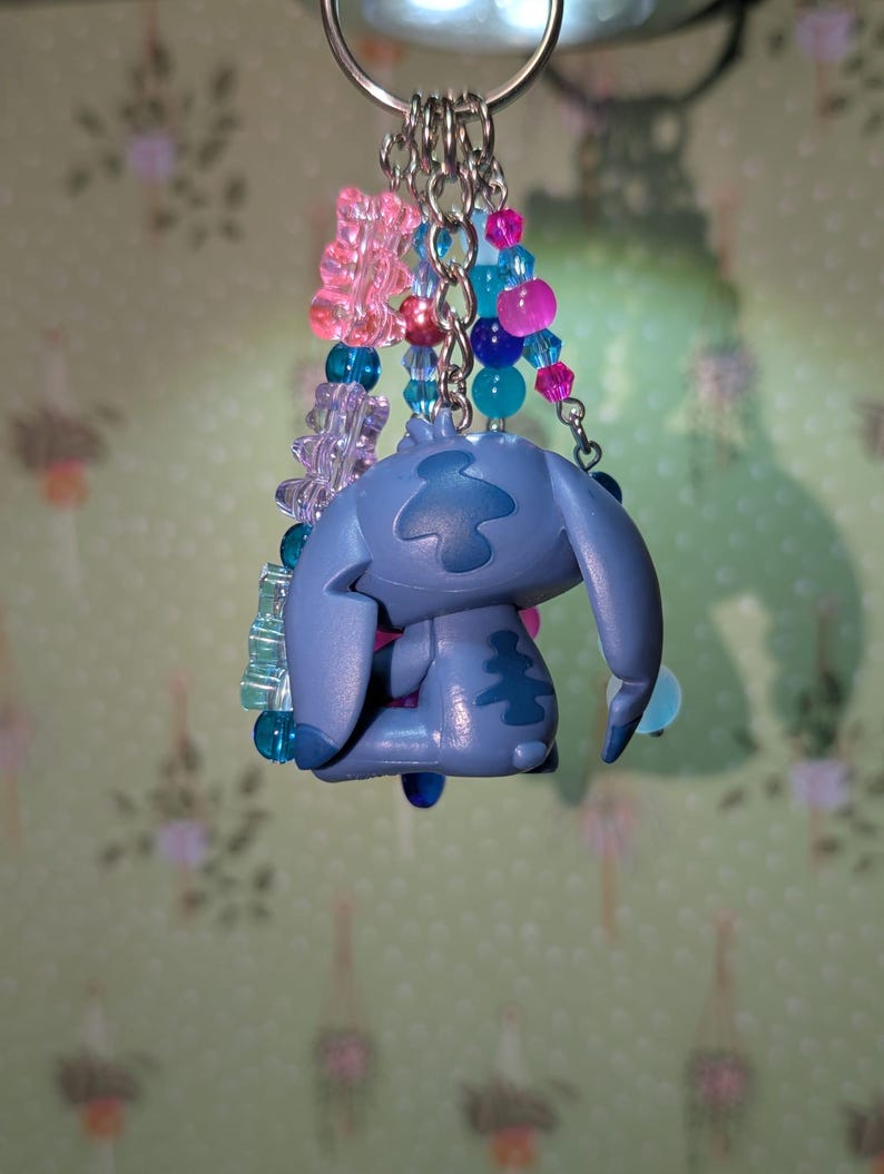 May include: A blue Stitch keychain with a silver keyring. The keychain features a Stitch figurine and colorful beads in pink, blue, and purple. The beads are strung together with silver chains.