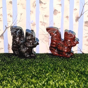 Puede incluir: Dos figuritas de ardilla talladas a mano, una negra y otra roja, hechas de piedra pulida. Las ardillas están sentadas sobre una superficie verde cubierta de hierba.