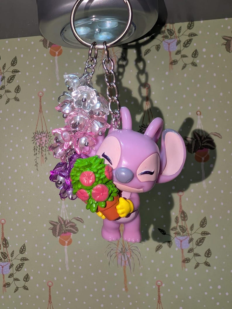 May include: A lavender-colored Angel keychain holding a potted plant with pink flowers and green leaves. The keychain also features a cluster of clear and pink beads. The keychain is attached to a silver metal ring.