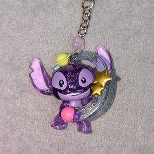 May include: A purple glittery Stitch keychain with a silver crescent moon and gold stars. The keychain has a silver chain and a pink bead. The character is holding a pink object and wearing a yellow hat.