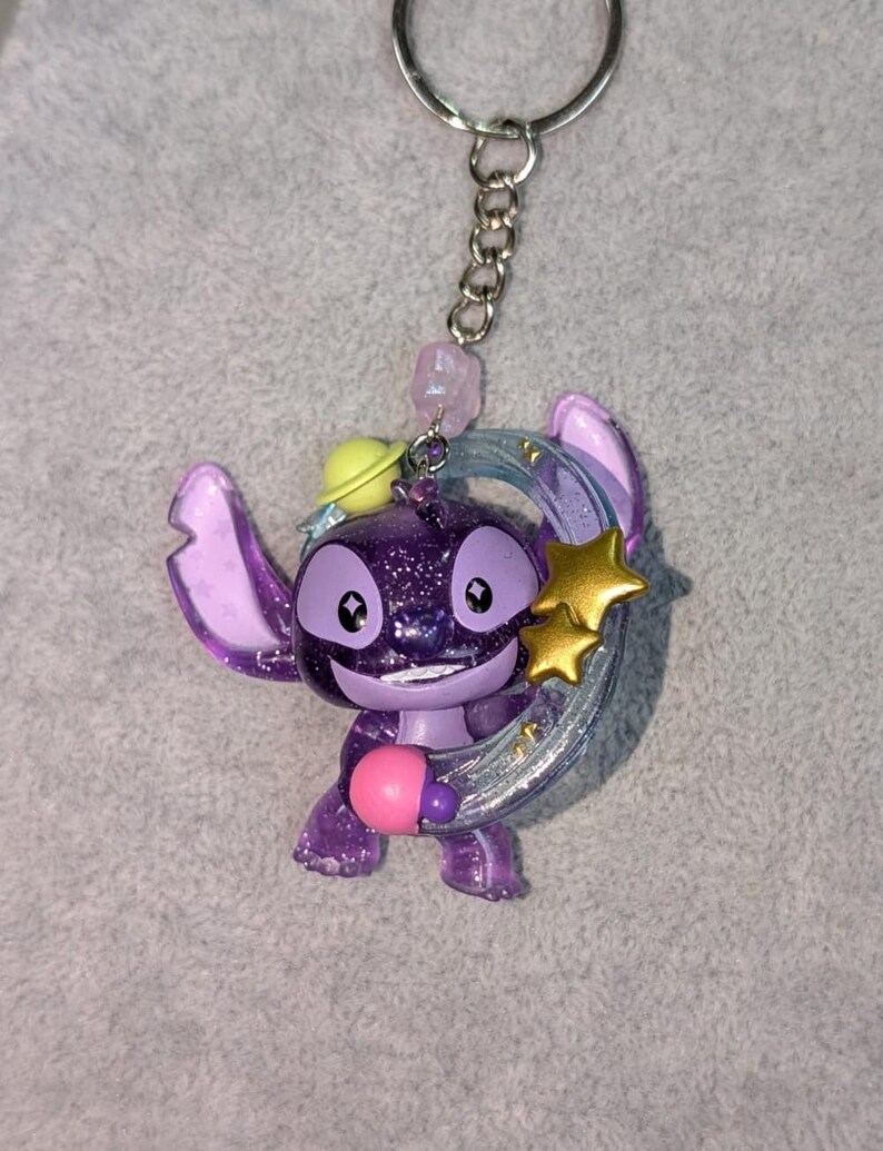 May include: A purple glittery Stitch keychain with a silver crescent moon and gold stars. The keychain has a silver chain and a pink bead. The character is holding a pink object and wearing a yellow hat.