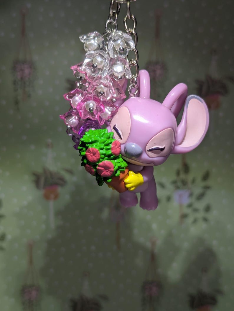 May include: A lavender-colored Angel keychain holding a small potted plant with pink flowers. The keychain features a silver chain and a cluster of clear and pink flower beads. The background is a light green wallpaper with hanging plant designs.