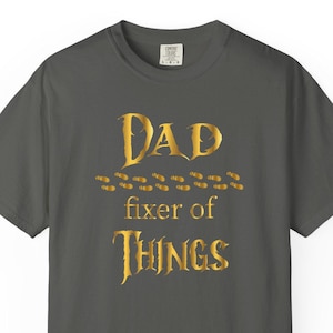 May include: Dark gray t-shirt with gold text that reads "Dad fixer of THINGS". The text is in a decorative font. The shirt is a short-sleeved crew neck.