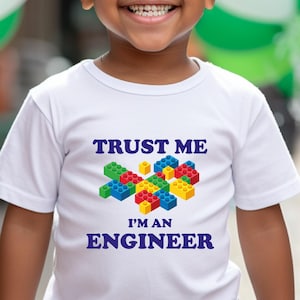 May include: A young child wearing a white t-shirt with the text "TRUST ME I'M AN ENGINEER" and a colorful block design. The child is also wearing a green cap and green pants, smiling in front of green and white balloons.