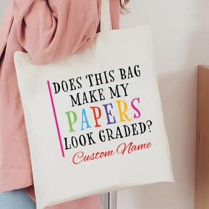 May include: Cream-colored tote bag with the text "DOES THIS BAG MAKE MY PAPERS LOOK GRADED? Custom Name" in colorful letters. The bag has a simple design with a rectangular shape and long straps.